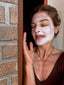 Woman Kim with a face mask applying it to her face, leaning against a brick wall.